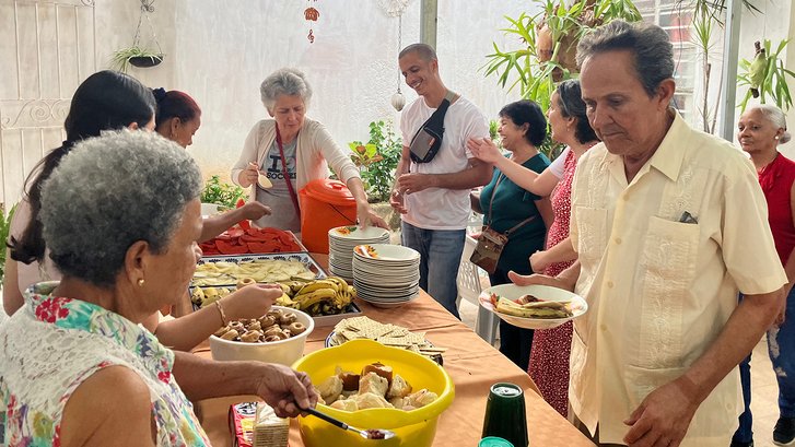 Eine Dame verteilt essen an Menschen verschiedensten Alters. Vor Ihr auf dem Tisch sind Obst, Keckse, Brot und verschiedenes weiteres Essen.