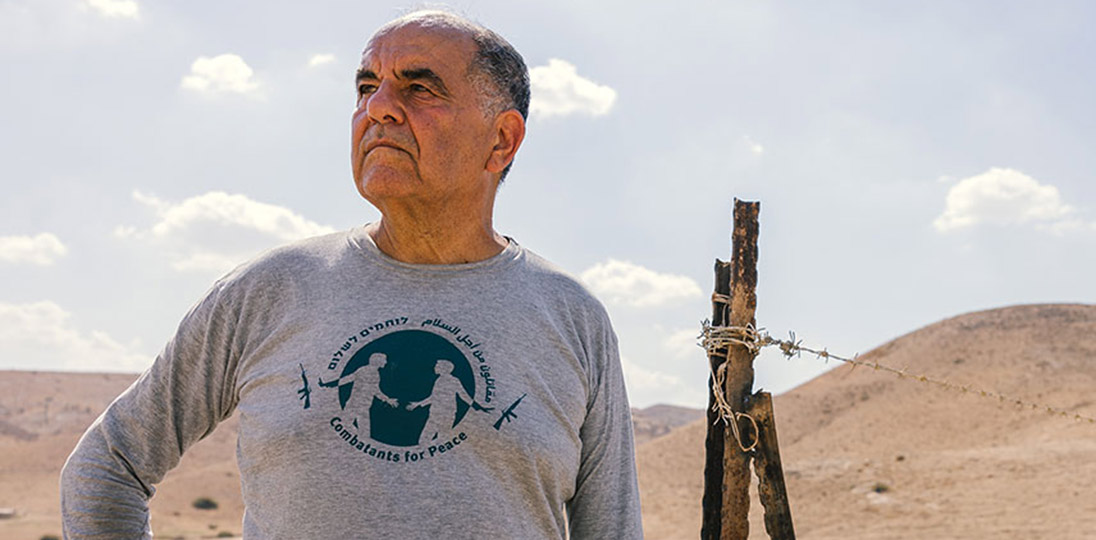 Elie Avidor von den Combatants for Peace, ein älterer Mann, steht in einer Wüstenlandschaft. Er trägt ein graues T-Shirt mit dem Logo der Organisation. 
